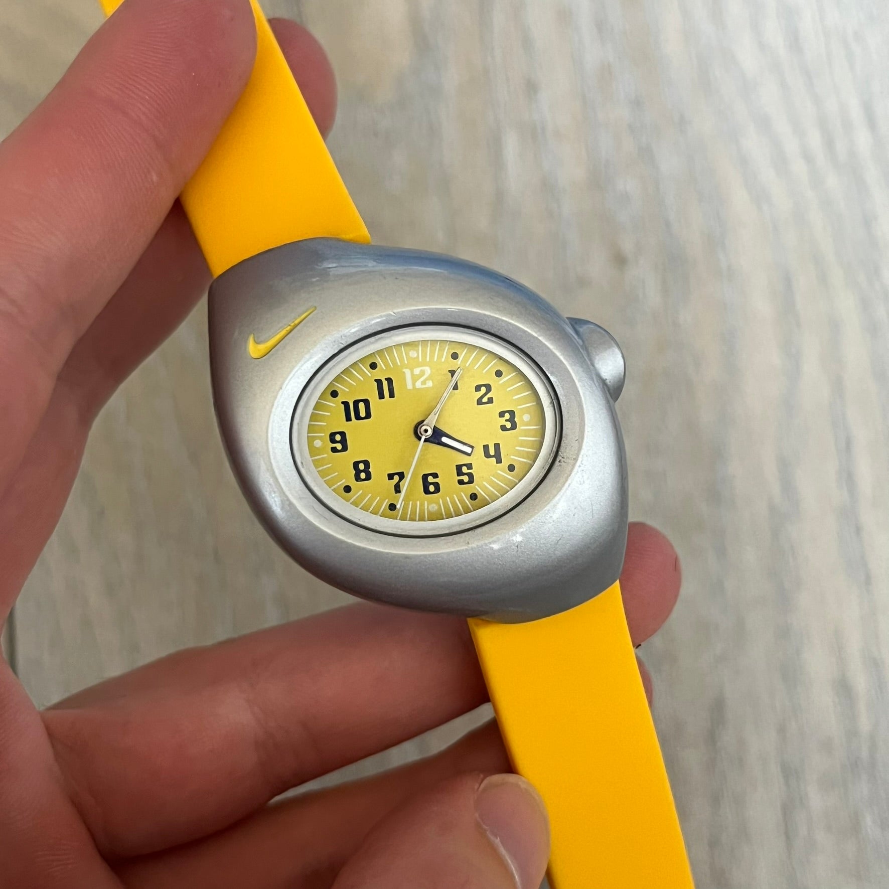 Nike Triax Junior WR0033 Yellow Analog Watch is a 2000s analog watch featuring a silver-tone case with a small yellow Nike logo above the dial. The yellow face is paired with white hands and white-and-black numerals for contrast. It is fitted with yellow replacement straps that are not original Nike straps. Constructed from stainless steel and plastic materials, the watch is water resistant up to 50 meters and includes US patent markings.