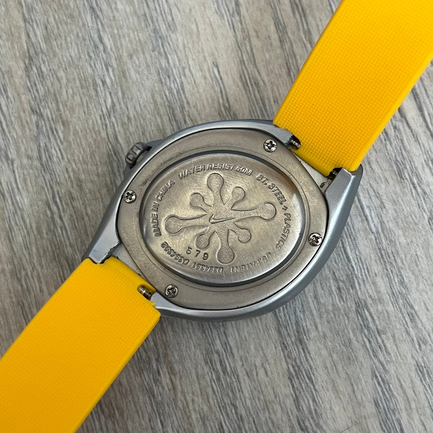 Nike Triax Junior WR0033 Yellow Analog Watch is a 2000s analog watch featuring a silver-tone case with a small yellow Nike logo above the dial. The yellow face is paired with white hands and white-and-black numerals for contrast. It is fitted with yellow replacement straps that are not original Nike straps. Constructed from stainless steel and plastic materials, the watch is water resistant up to 50 meters and includes US patent markings.