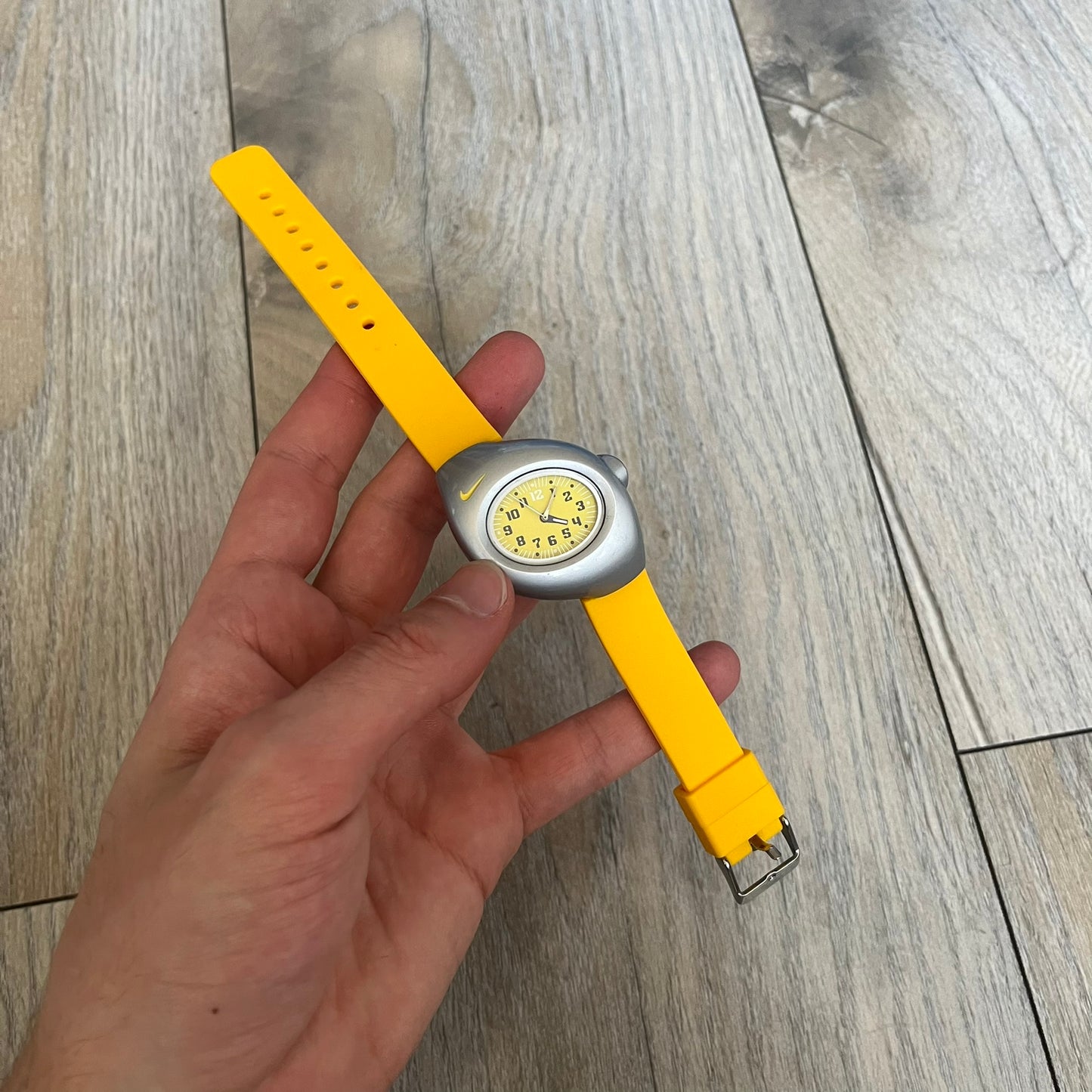 Nike Triax Junior WR0033 Yellow Analog Watch is a 2000s analog watch featuring a silver-tone case with a small yellow Nike logo above the dial. The yellow face is paired with white hands and white-and-black numerals for contrast. It is fitted with yellow replacement straps that are not original Nike straps. Constructed from stainless steel and plastic materials, the watch is water resistant up to 50 meters and includes US patent markings.