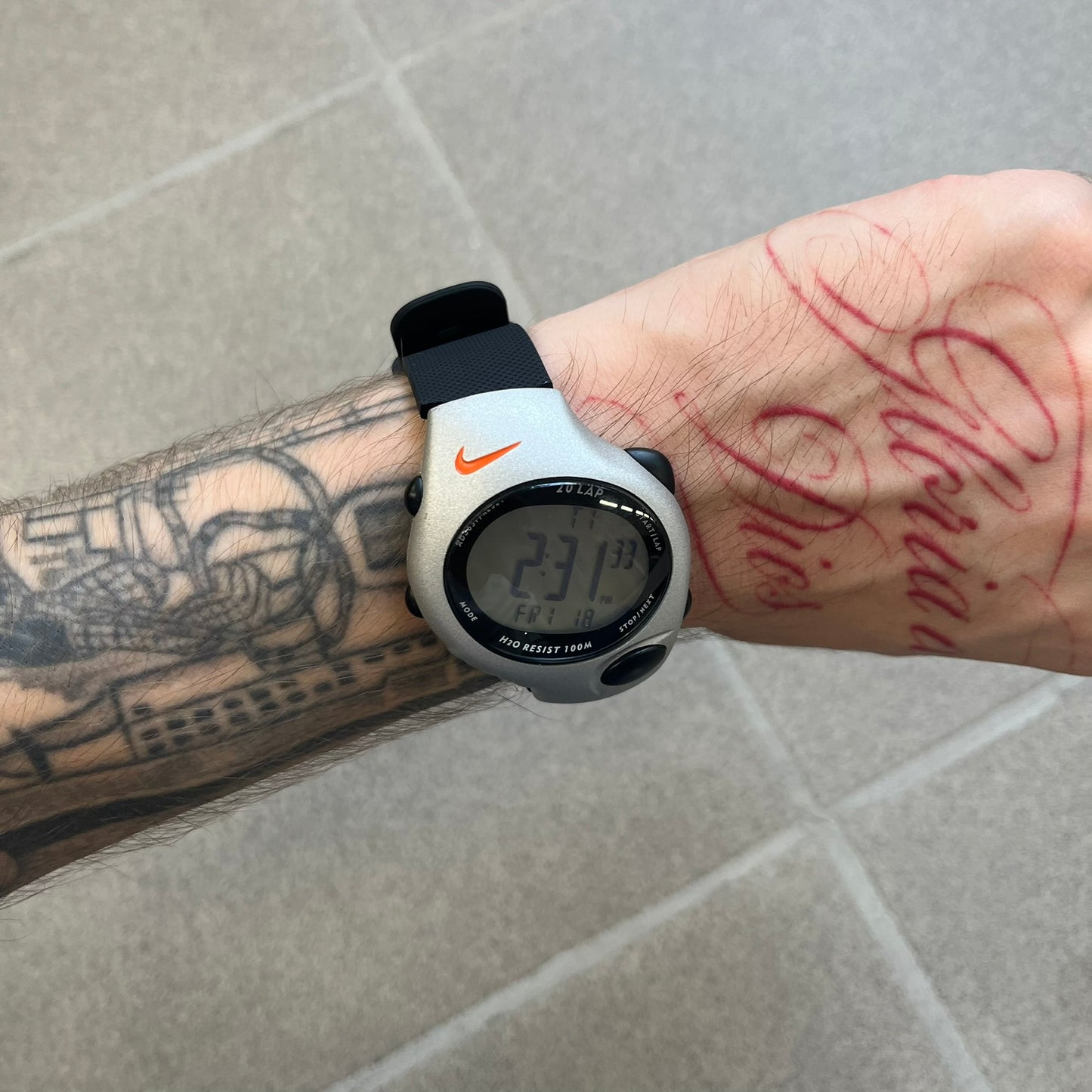 Nike Triax Regular 20 Silver Analog Watch features a silver-tone case with four black side buttons and a front button for the backlight function. The watch has an orange Nike Swoosh on the top of the case, 20-lap memory, multiple modes, and 100m water resistance. It comes with black replacement straps that are not original Nike.
