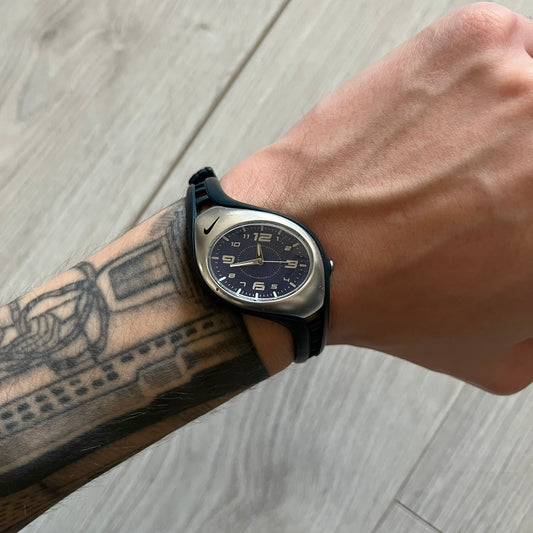 Nike Triax Roar Junior WK0007 analog watch featuring a navy rubber strap with matching navy case sides and a silver-tone front case. The metallic blue dial has a high-shine finish, silver hour markers, and silver hands for clear visibility. Built with stainless steel and metal components, the watch is water resistant up to 50 meters