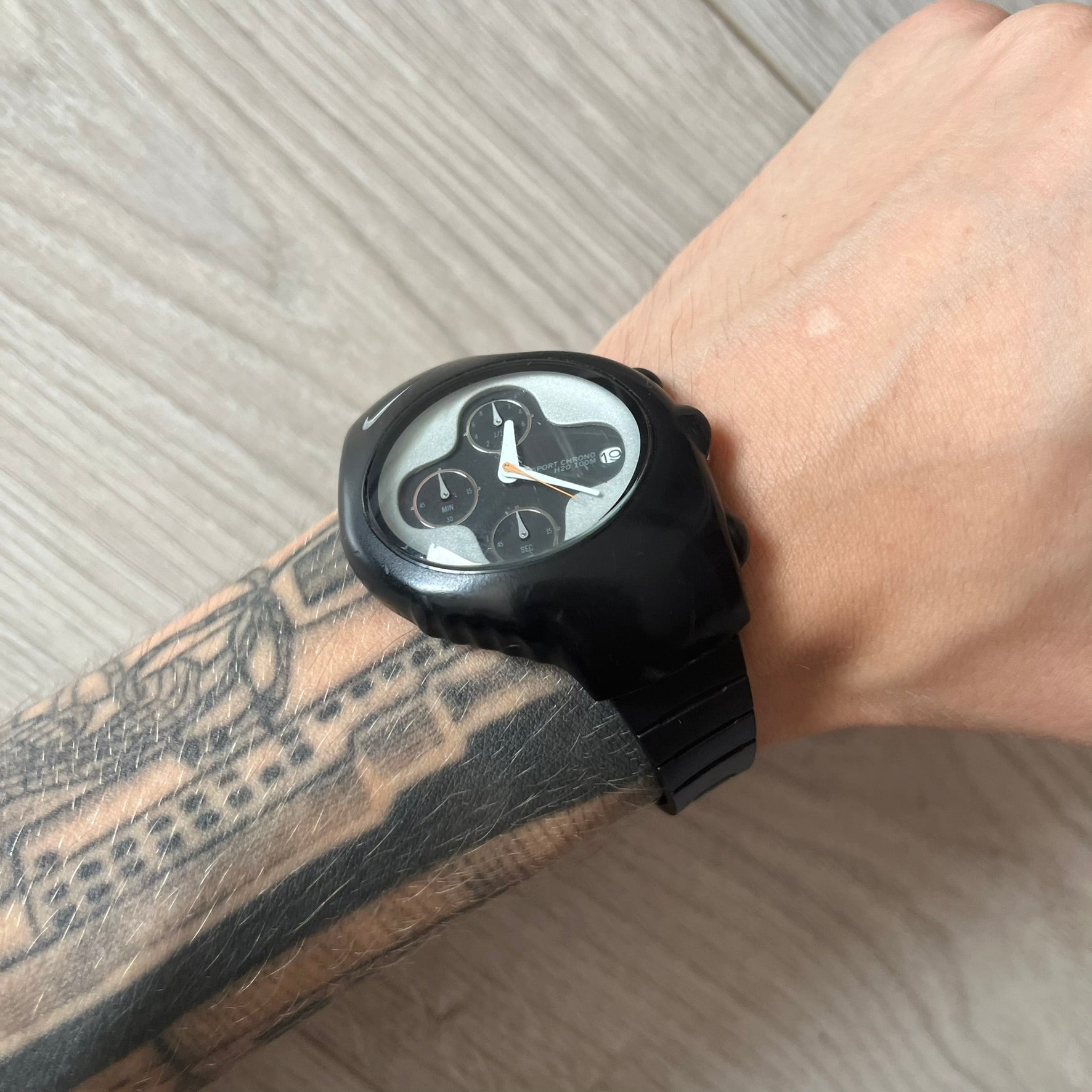 Black smartwatch on a wrist with a tattooed arm, on a light wooden surface.