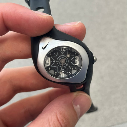 Nike Triax Sweeper is a really small analog watch that features a compact silver-tone case with a black Nike Swoosh at the top. It has black rubber straps and a black dial with white hour markers and decorative star and dot graphics. The watch is water resistant to 50 metres.