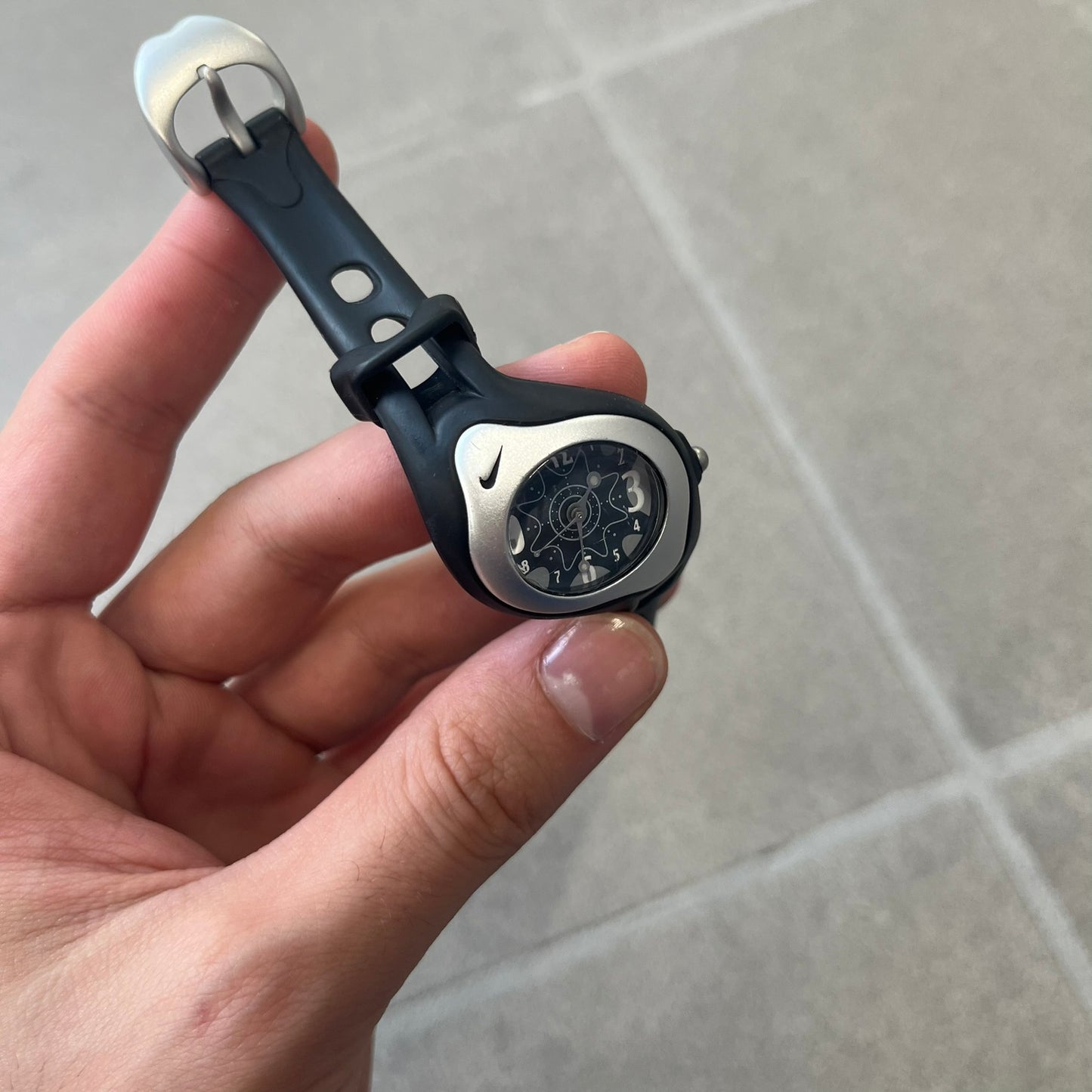 Nike Triax Sweeper is a really small analog watch that features a compact silver-tone case with a black Nike Swoosh at the top. It has black rubber straps and a black dial with white hour markers and decorative star and dot graphics. The watch is water resistant to 50 metres.