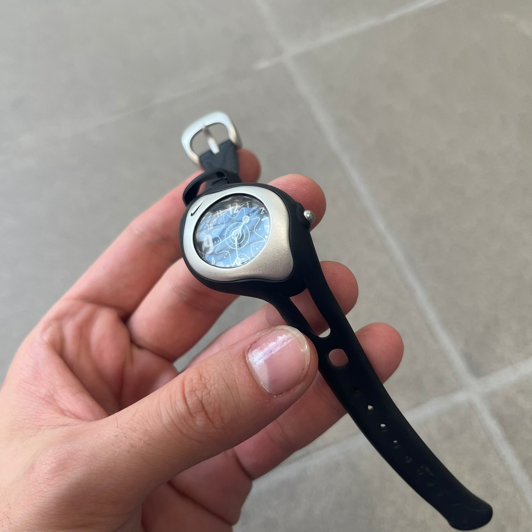 Nike Triax Sweeper is a really small analog watch that features a compact silver-tone case with a black Nike Swoosh at the top. It has black rubber straps and a black dial with white hour markers and decorative star and dot graphics. The watch is water resistant to 50 metres.