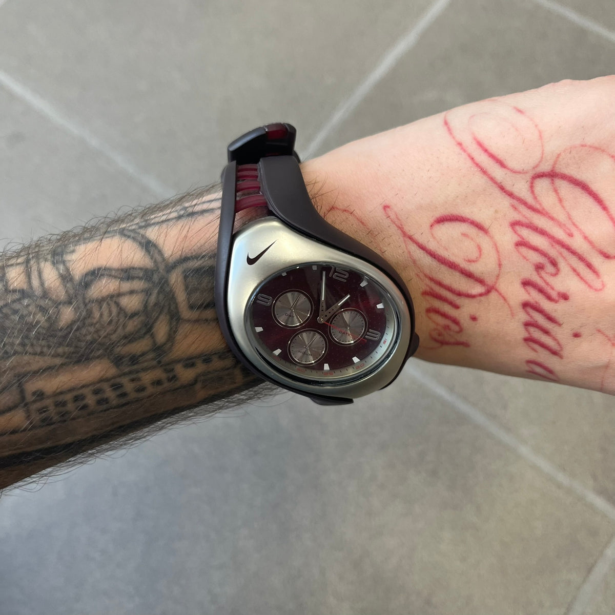 Nike Triax Swift 3i Burgundy WR0091 Analog Watch comes with burgundy and red rubber straps and a silver case featuring a dark Nike Swoosh. The burgundy metallic dial has a dotted pattern, three silver subdials for day, date, and hour and silver hands. Seconds markings are printed on the inner edge of the case and are visible only at an angle.