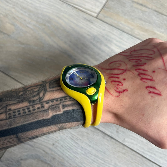 Nike Triax Swift Brazil WR0078 Analog Watch is an analog sports watch featuring a yellow rubber strap integrated into the case design, forming a one-piece construction. The case is metal green with a yellow Nike Swoosh at the top and a yellow side button used to activate the backlight. The dial is blue and white with the Brazil national football team crest and “Brazil” text, paired with yellow hands. It is water resistant up to 100 meters.