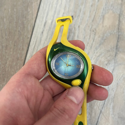 Nike Triax Swift Brazil WR0078 Analog Watch is an analog sports watch featuring a yellow rubber strap integrated into the case design, forming a one-piece construction. The case is metal green with a yellow Nike Swoosh at the top and a yellow side button used to activate the backlight. The dial is blue and white with the Brazil national football team crest and “Brazil” text, paired with yellow hands. It is water resistant up to 100 meters.