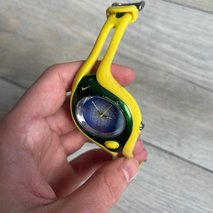 Nike Triax Swift Brazil WR0078 Analog Watch is an analog sports watch featuring a yellow rubber strap integrated into the case design, forming a one-piece construction. The case is metal green with a yellow Nike Swoosh at the top and a yellow side button used to activate the backlight. The dial is blue and white with the Brazil national football team crest and “Brazil” text, paired with yellow hands. It is water resistant up to 100 meters.