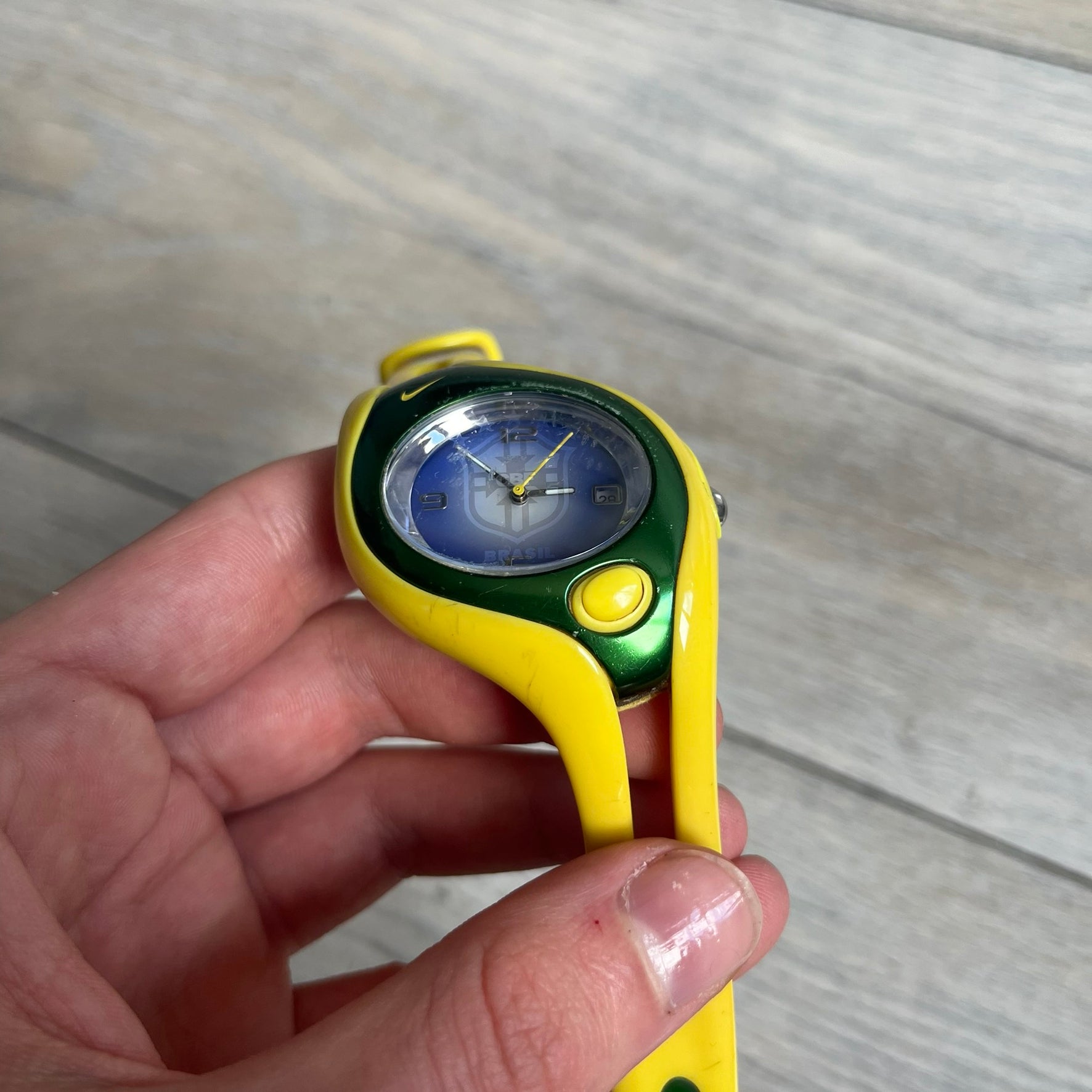 Nike Triax Swift Brazil WR0078 Analog Watch is an analog sports watch featuring a yellow rubber strap integrated into the case design, forming a one-piece construction. The case is metal green with a yellow Nike Swoosh at the top and a yellow side button used to activate the backlight. The dial is blue and white with the Brazil national football team crest and “Brazil” text, paired with yellow hands. It is water resistant up to 100 meters.