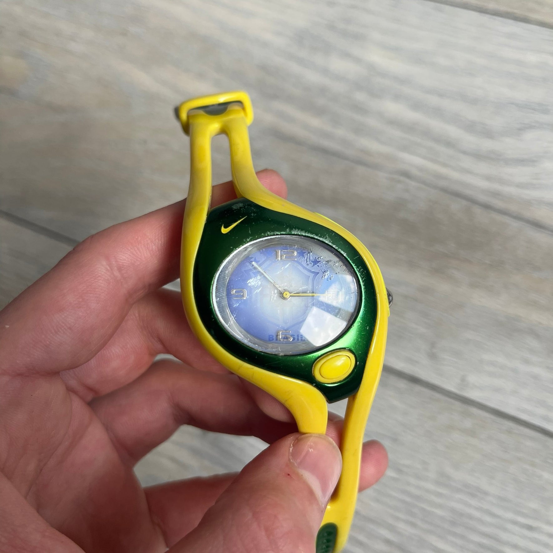 Nike Triax Swift Brazil WR0078 Analog Watch is an analog sports watch featuring a yellow rubber strap integrated into the case design, forming a one-piece construction. The case is metal green with a yellow Nike Swoosh at the top and a yellow side button used to activate the backlight. The dial is blue and white with the Brazil national football team crest and “Brazil” text, paired with yellow hands. It is water resistant up to 100 meters.