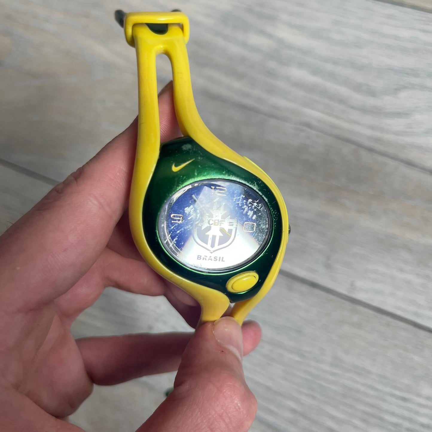Nike Triax Swift Brazil WR0078 Analog Watch is an analog sports watch featuring a yellow rubber strap integrated into the case design, forming a one-piece construction. The case is metal green with a yellow Nike Swoosh at the top and a yellow side button used to activate the backlight. The dial is blue and white with the Brazil national football team crest and “Brazil” text, paired with yellow hands. It is water resistant up to 100 meters.