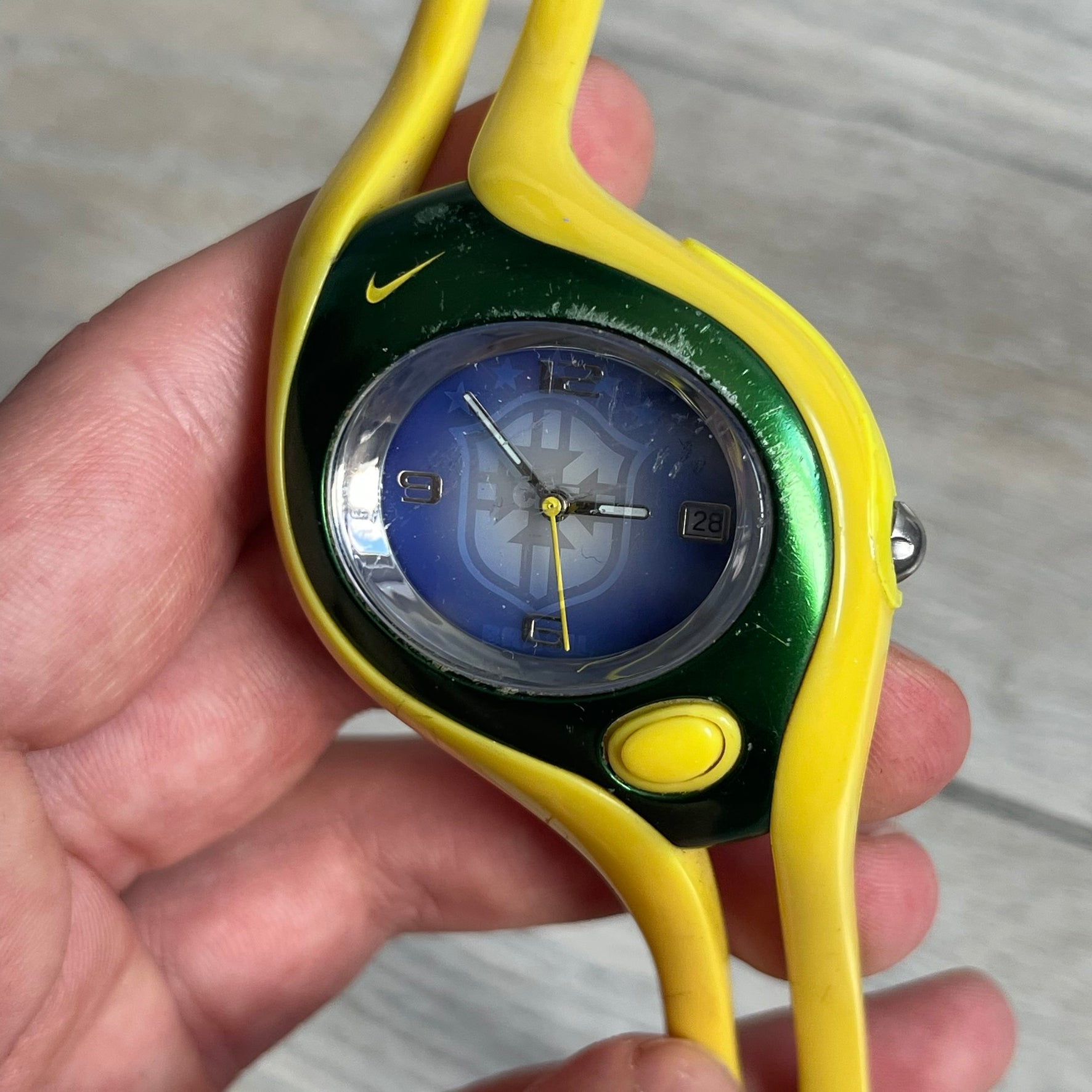 Nike Triax Swift Brazil WR0078 Analog Watch is an analog sports watch featuring a yellow rubber strap integrated into the case design, forming a one-piece construction. The case is metal green with a yellow Nike Swoosh at the top and a yellow side button used to activate the backlight. The dial is blue and white with the Brazil national football team crest and “Brazil” text, paired with yellow hands. It is water resistant up to 100 meters.