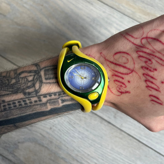 Nike Triax Swift Brazil WR0078 Analog Watch is an analog sports watch featuring a yellow rubber strap integrated into the case design, forming a one-piece construction. The case is metal green with a yellow Nike Swoosh at the top and a yellow side button used to activate the backlight. The dial is blue and white with the Brazil national football team crest and “Brazil” text, paired with yellow hands. It is water resistant up to 100 meters.