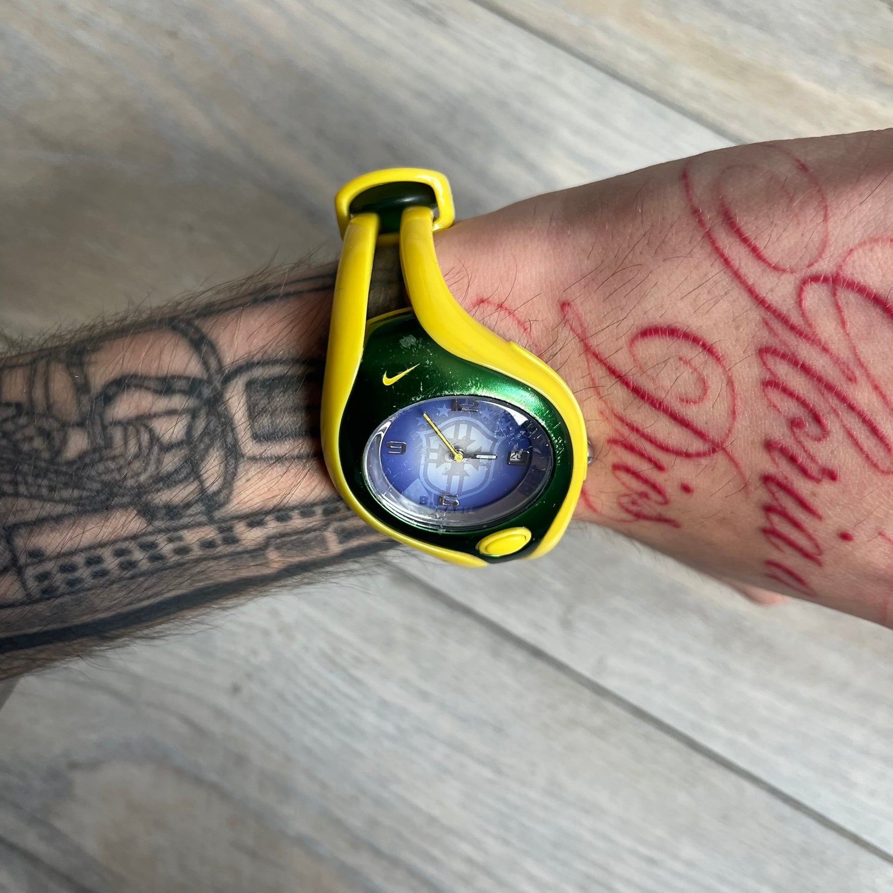 Nike Triax Swift Brazil WR0078 Analog Watch is an analog sports watch featuring a yellow rubber strap integrated into the case design, forming a one-piece construction. The case is metal green with a yellow Nike Swoosh at the top and a yellow side button used to activate the backlight. The dial is blue and white with the Brazil national football team crest and “Brazil” text, paired with yellow hands. It is water resistant up to 100 meters.