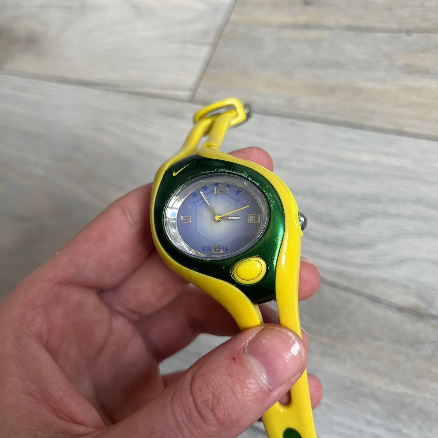 Nike Triax Swift Brazil WR0078 Analog Watch is an analog sports watch featuring a yellow rubber strap integrated into the case design, forming a one-piece construction. The case is metal green with a yellow Nike Swoosh at the top and a yellow side button used to activate the backlight. The dial is blue and white with the Brazil national football team crest and “Brazil” text, paired with yellow hands. It is water resistant up to 100 meters.