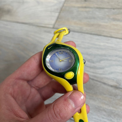 Nike Triax Swift Brazil WR0078 Analog Watch is an analog sports watch featuring a yellow rubber strap integrated into the case design, forming a one-piece construction. The case is metal green with a yellow Nike Swoosh at the top and a yellow side button used to activate the backlight. The dial is blue and white with the Brazil national football team crest and “Brazil” text, paired with yellow hands. It is water resistant up to 100 meters.