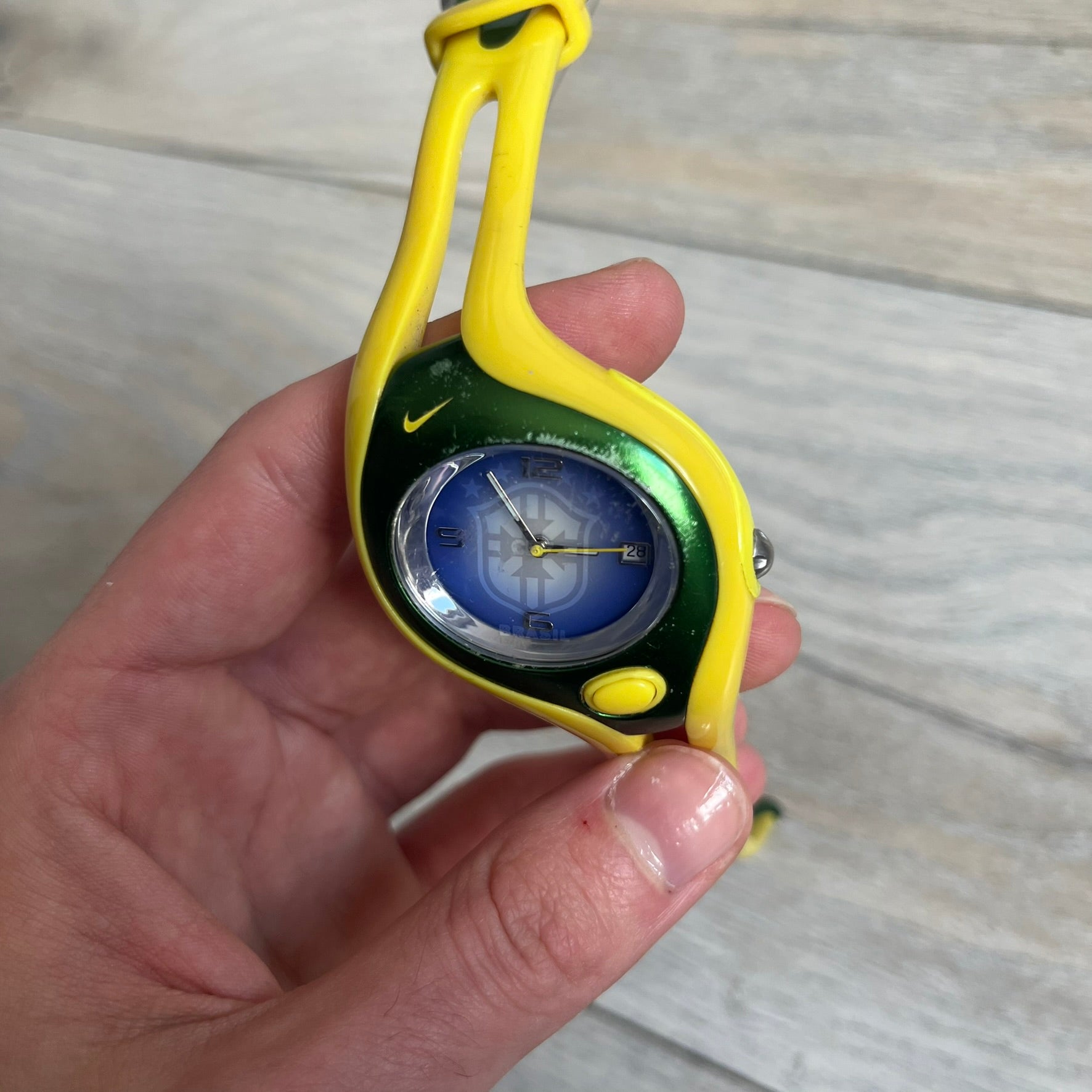 Nike Triax Swift Brazil WR0078 Analog Watch is an analog sports watch featuring a yellow rubber strap integrated into the case design, forming a one-piece construction. The case is metal green with a yellow Nike Swoosh at the top and a yellow side button used to activate the backlight. The dial is blue and white with the Brazil national football team crest and “Brazil” text, paired with yellow hands. It is water resistant up to 100 meters.