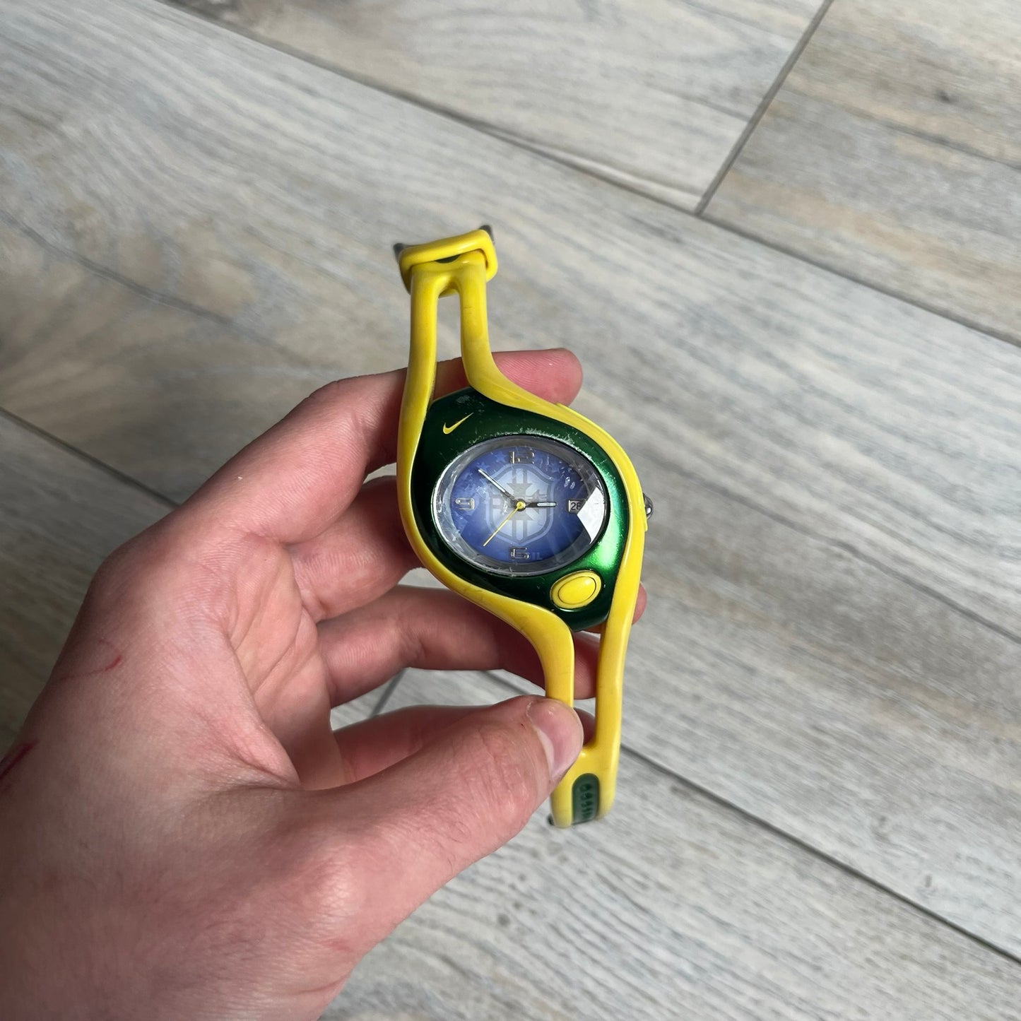Nike Triax Swift Brazil WR0078 Analog Watch is an analog sports watch featuring a yellow rubber strap integrated into the case design, forming a one-piece construction. The case is metal green with a yellow Nike Swoosh at the top and a yellow side button used to activate the backlight. The dial is blue and white with the Brazil national football team crest and “Brazil” text, paired with yellow hands. It is water resistant up to 100 meters.