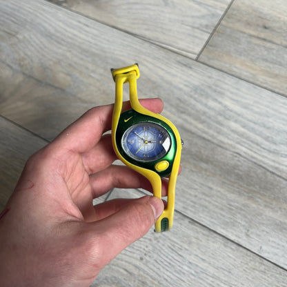 Nike Triax Swift Brazil WR0078 Analog Watch is an analog sports watch featuring a yellow rubber strap integrated into the case design, forming a one-piece construction. The case is metal green with a yellow Nike Swoosh at the top and a yellow side button used to activate the backlight. The dial is blue and white with the Brazil national football team crest and “Brazil” text, paired with yellow hands. It is water resistant up to 100 meters.