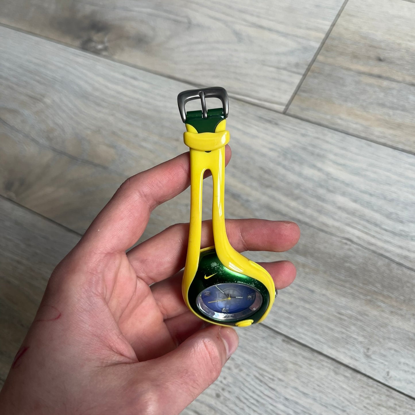 Nike Triax Swift Brazil WR0078 Analog Watch is an analog sports watch featuring a yellow rubber strap integrated into the case design, forming a one-piece construction. The case is metal green with a yellow Nike Swoosh at the top and a yellow side button used to activate the backlight. The dial is blue and white with the Brazil national football team crest and “Brazil” text, paired with yellow hands. It is water resistant up to 100 meters.