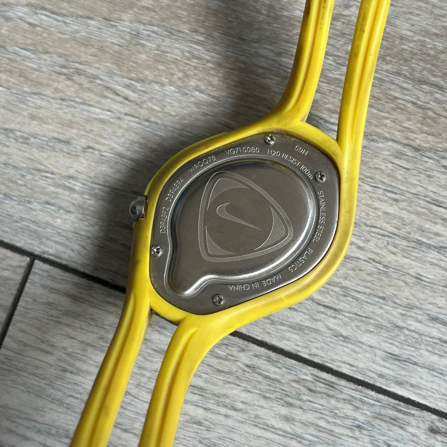 Nike Triax Swift Brazil WR0078 Analog Watch is an analog sports watch featuring a yellow rubber strap integrated into the case design, forming a one-piece construction. The case is metal green with a yellow Nike Swoosh at the top and a yellow side button used to activate the backlight. The dial is blue and white with the Brazil national football team crest and “Brazil” text, paired with yellow hands. It is water resistant up to 100 meters.