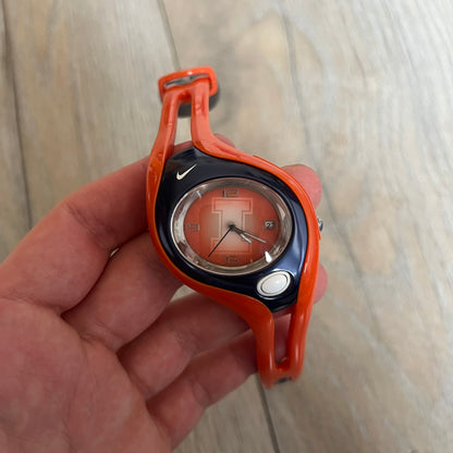 Nike Triax Swift Illinois University WR0078 Analog Watch is a sports watch featuring an integrated one-piece construction with orange straps and navy accents. The dark navy stainless steel case includes a white Nike Swoosh and a white button that activates the light blue backlight. The orange dial features the large Illinois University “I” logo. Made from stainless steel and plastic materials, the watch is water resistant up to 100 meters.