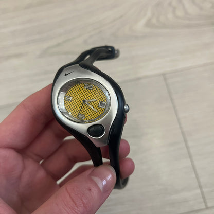 Nike Triax Swift WR0078 analog wristwatch with black rubber straps and a matching rubber outer case. The internal case is silver-tone metal, fitted with a black side button and black Nike Swoosh branding. Features a yellow dotted dial with silver hands and markers, paired with an integrated turquoise backlight that illuminates the face when activated. Sport-oriented construction with a clean, functional layout typical of early-2000s Nike Triax models.&nbsp;