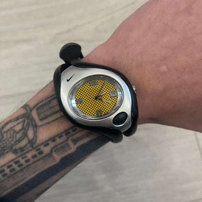 Nike Triax Swift WR0078 analog wristwatch with black rubber straps and a matching rubber outer case. The internal case is silver-tone metal, fitted with a black side button and black Nike Swoosh branding. Features a yellow dotted dial with silver hands and markers, paired with an integrated turquoise backlight that illuminates the face when activated. Sport-oriented construction with a clean, functional layout typical of early-2000s Nike Triax models.&nbsp;