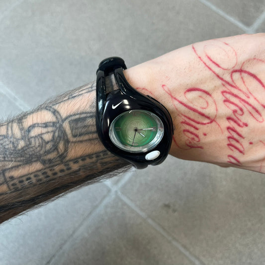 Nike Triax Swift 'Celtic F.C.' WR0078 Black Analog Watch featuring black straps and a matching black outer shell covering the metal case. The inner case is metallic black with a white light button and a Nike Swoosh on the top. The dial has a light green gradient finish with a large Celtic F.C. crest in the center. Analog display with an integrated backlight that illuminates the dial.