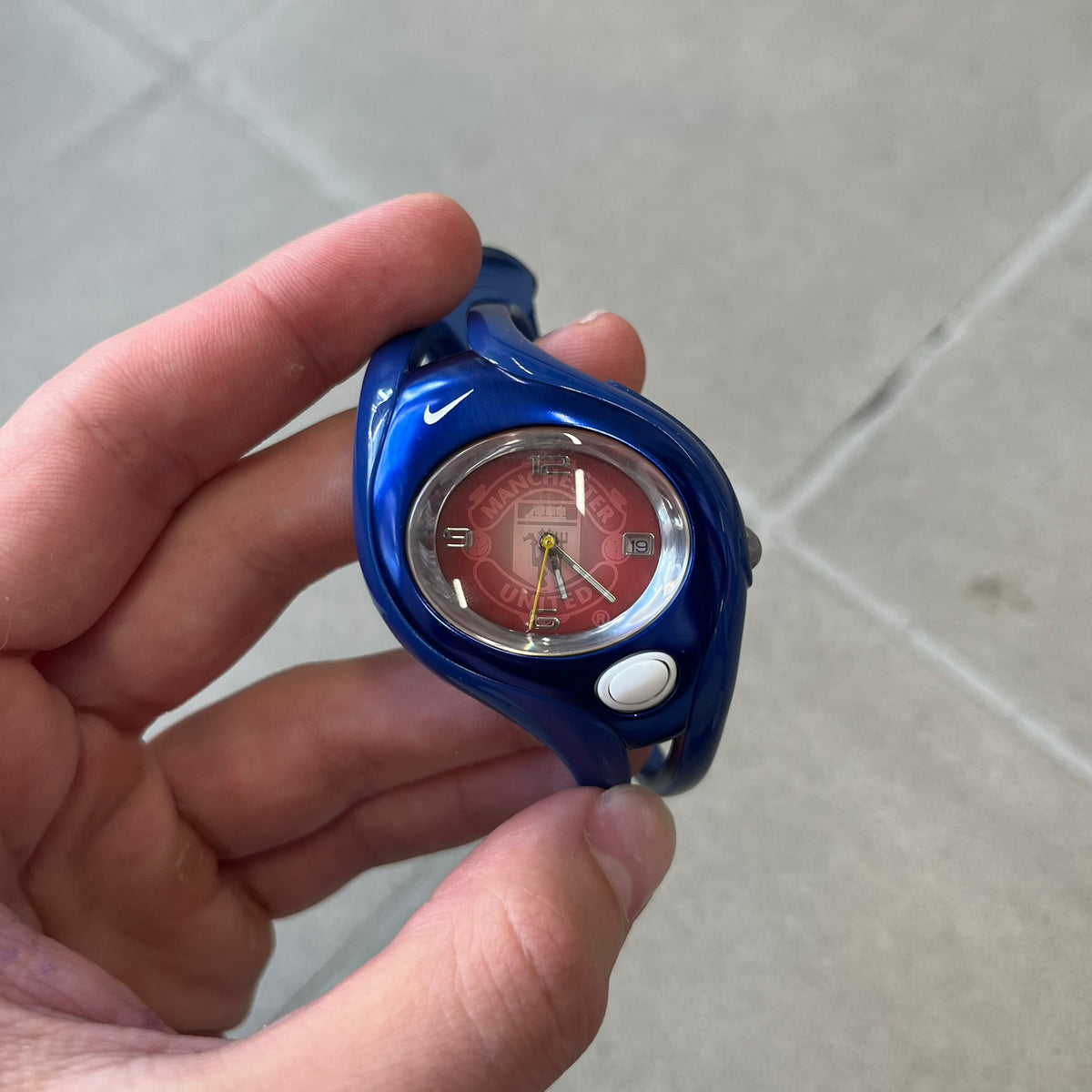 Nike Triax Swift 'Manchester United' WR0078 Blue Analog Watch featuring blue straps and a matching blue outer shell covering the metal case. The inner case is metallic blue with a white light button and a Nike Swoosh on the top. The dial has a light red gradient finish with a large Manchester United crest in the center. Analog display with an integrated backlight that illuminates the dial.