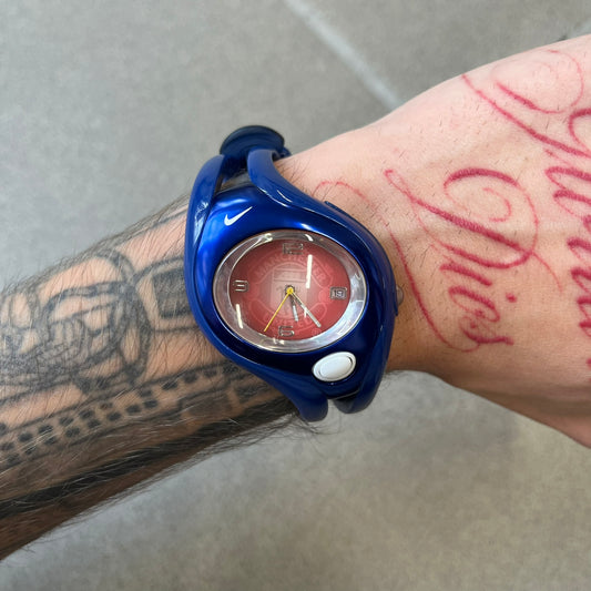 Nike Triax Swift 'Manchester United' WR0078 Blue Analog Watch featuring blue straps and a matching blue outer shell covering the metal case. The inner case is metallic blue with a white light button and a Nike Swoosh on the top. The dial has a light red gradient finish with a large Manchester United crest in the center. Analog display with an integrated backlight that illuminates the dial.