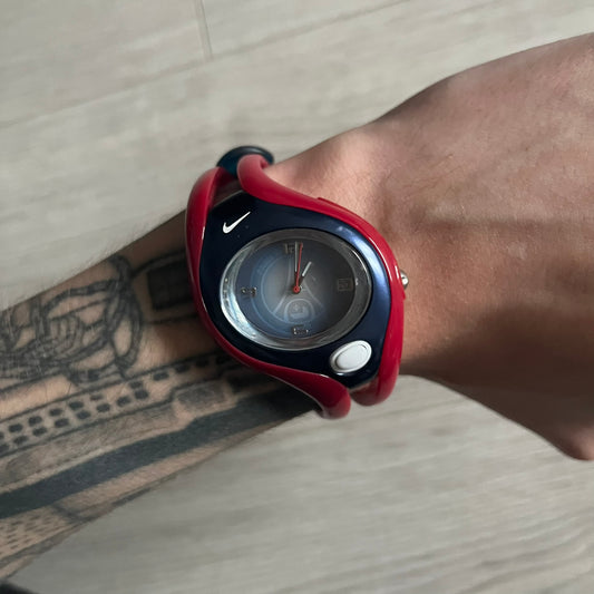 Nike Triax Swift “PSG” WR0078 Analog Watch featuring red rubberized straps and a matching red outer case shell covering the metal housing. The inner case is navy with a white light button and a Nike Swoosh on the side. The dial has a white-to-blue mirrored gradient finish with a large Paris Saint-Germain crest centered on the face, visible depending on light angle. Analog display with an integrated turquoise backlight that illuminates the dial. Designed as an official Nike Triax Swift collaboration tied to 