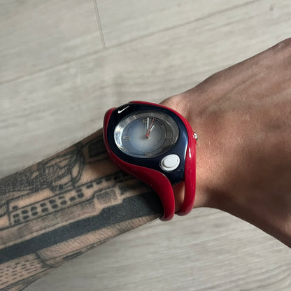 Nike Triax Swift “PSG” WR0078 Analog Watch featuring red rubberized straps and a matching red outer case shell covering the metal housing. The inner case is navy with a white light button and a Nike Swoosh on the side. The dial has a white-to-blue mirrored gradient finish with a large Paris Saint-Germain crest centered on the face, visible depending on light angle. Analog display with an integrated turquoise backlight that illuminates the dial. Designed as an official Nike Triax Swift collaboration tied to 