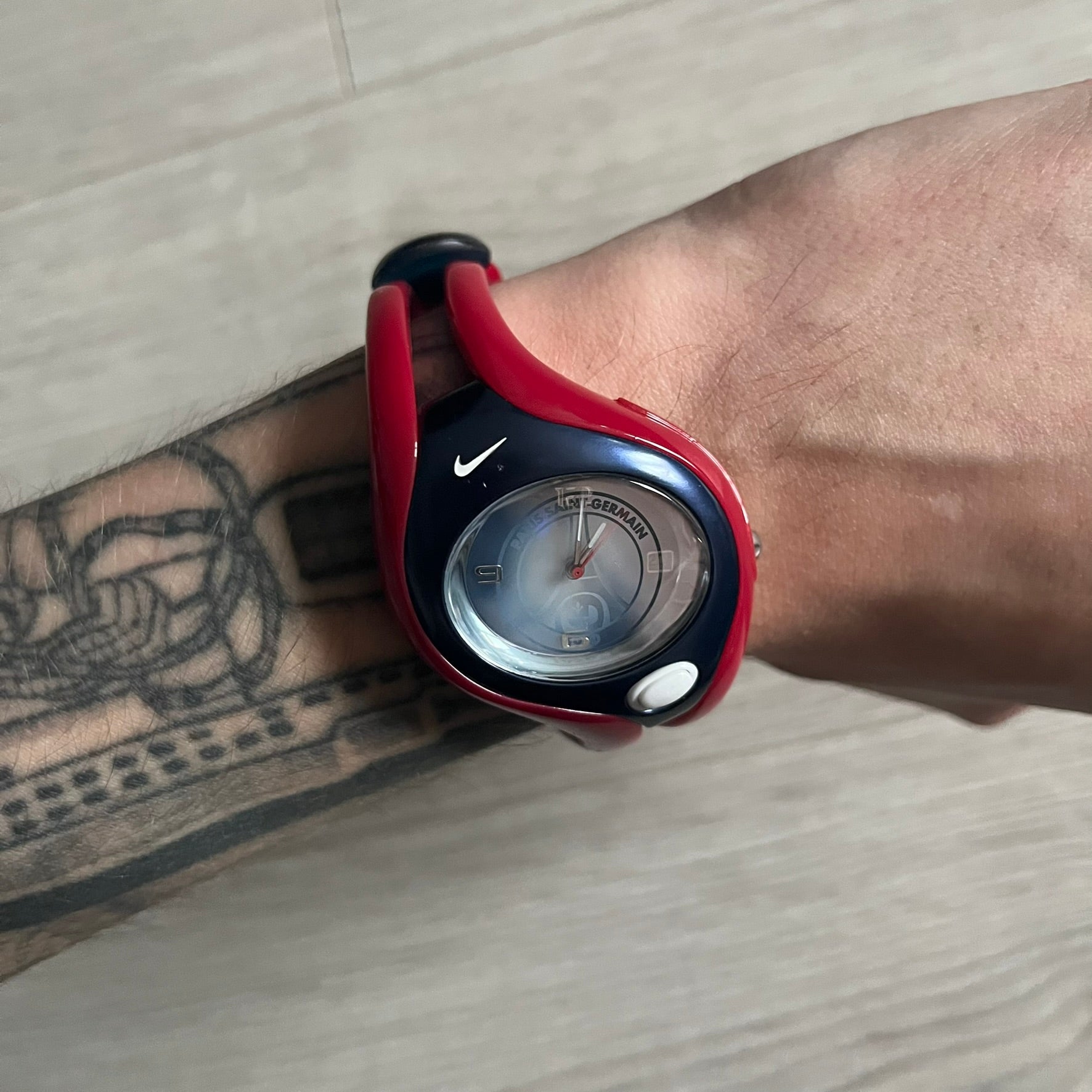 Nike Triax Swift “PSG” WR0078 Analog Watch featuring red rubberized straps and a matching red outer case shell covering the metal housing. The inner case is navy with a white light button and a Nike Swoosh on the side. The dial has a white-to-blue mirrored gradient finish with a large Paris Saint-Germain crest centered on the face, visible depending on light angle. Analog display with an integrated turquoise backlight that illuminates the dial. Designed as an official Nike Triax Swift collaboration tied to 