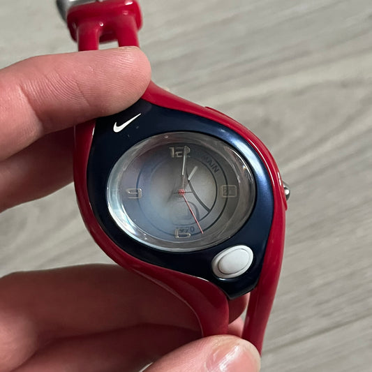 Nike Triax Swift “PSG” WR0078 Analog Watch featuring red rubberized straps and a matching red outer case shell covering the metal housing. The inner case is navy with a white light button and a Nike Swoosh on the side. The dial has a white-to-blue mirrored gradient finish with a large Paris Saint-Germain crest centered on the face, visible depending on light angle. Analog display with an integrated turquoise backlight that illuminates the dial. Designed as an official Nike Triax Swift collaboration tied to 