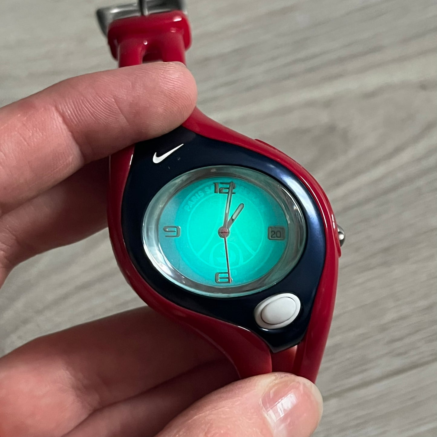 Nike Triax Swift “PSG” WR0078 Analog Watch featuring red rubberized straps and a matching red outer case shell covering the metal housing. The inner case is navy with a white light button and a Nike Swoosh on the side. The dial has a white-to-blue mirrored gradient finish with a large Paris Saint-Germain crest centered on the face, visible depending on light angle. Analog display with an integrated turquoise backlight that illuminates the dial. Designed as an official Nike Triax Swift collaboration tied to 