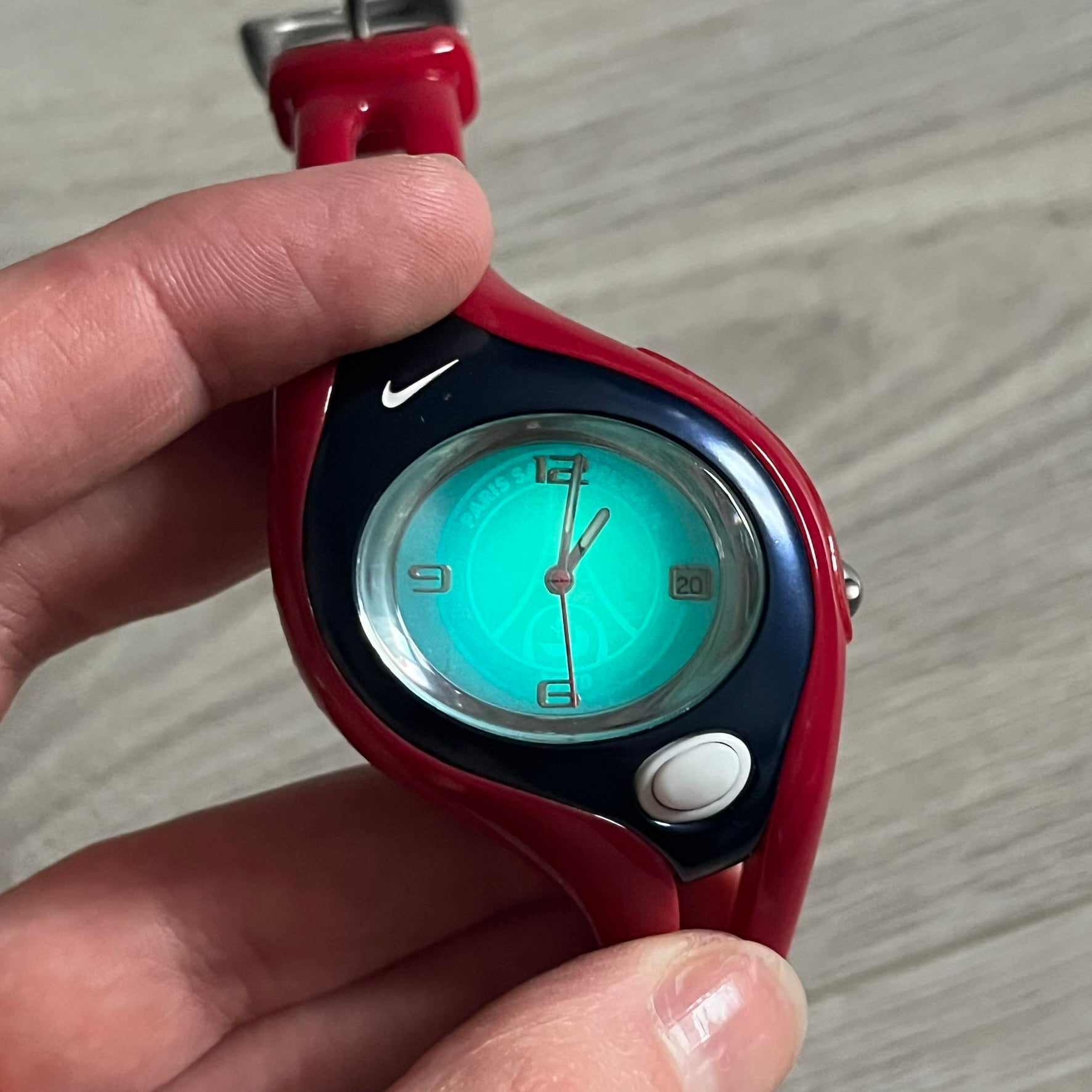 Nike Triax Swift “PSG” WR0078 Analog Watch featuring red rubberized straps and a matching red outer case shell covering the metal housing. The inner case is navy with a white light button and a Nike Swoosh on the side. The dial has a white-to-blue mirrored gradient finish with a large Paris Saint-Germain crest centered on the face, visible depending on light angle. Analog display with an integrated turquoise backlight that illuminates the dial. Designed as an official Nike Triax Swift collaboration tied to 
