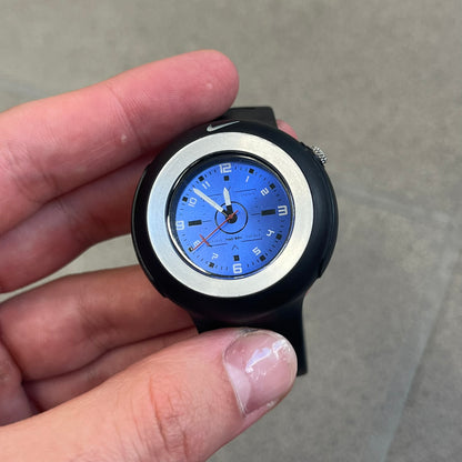 Nike Triax WK0009 Timing Range Blue Analog Watch features a black rubber strap and black rubber case with a silver-tone outline around the dial. The case has a grey Nike Swoosh at the top, while the metallic blue dial features white numerals and black hour markers.
