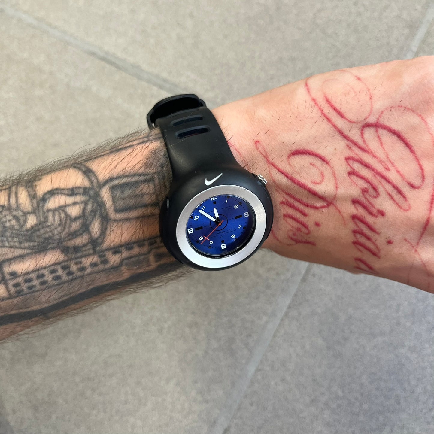Nike Triax WK0009 Timing Range Blue Analog Watch features a black rubber strap and black rubber case with a silver-tone outline around the dial. The case has a grey Nike Swoosh at the top, while the metallic blue dial features white numerals and black hour markers.
