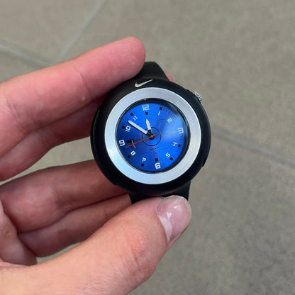 Nike Triax WK0009 Timing Range Blue Analog Watch features a black rubber strap and black rubber case with a silver-tone outline around the dial. The case has a grey Nike Swoosh at the top, while the metallic blue dial features white numerals and black hour markers.
