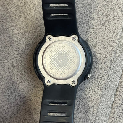 Nike Triax WK0009 Timing Range Blue Analog Watch features a black rubber strap and black rubber case with a silver-tone outline around the dial. The case has a grey Nike Swoosh at the top, while the metallic blue dial features white numerals and black hour markers.
