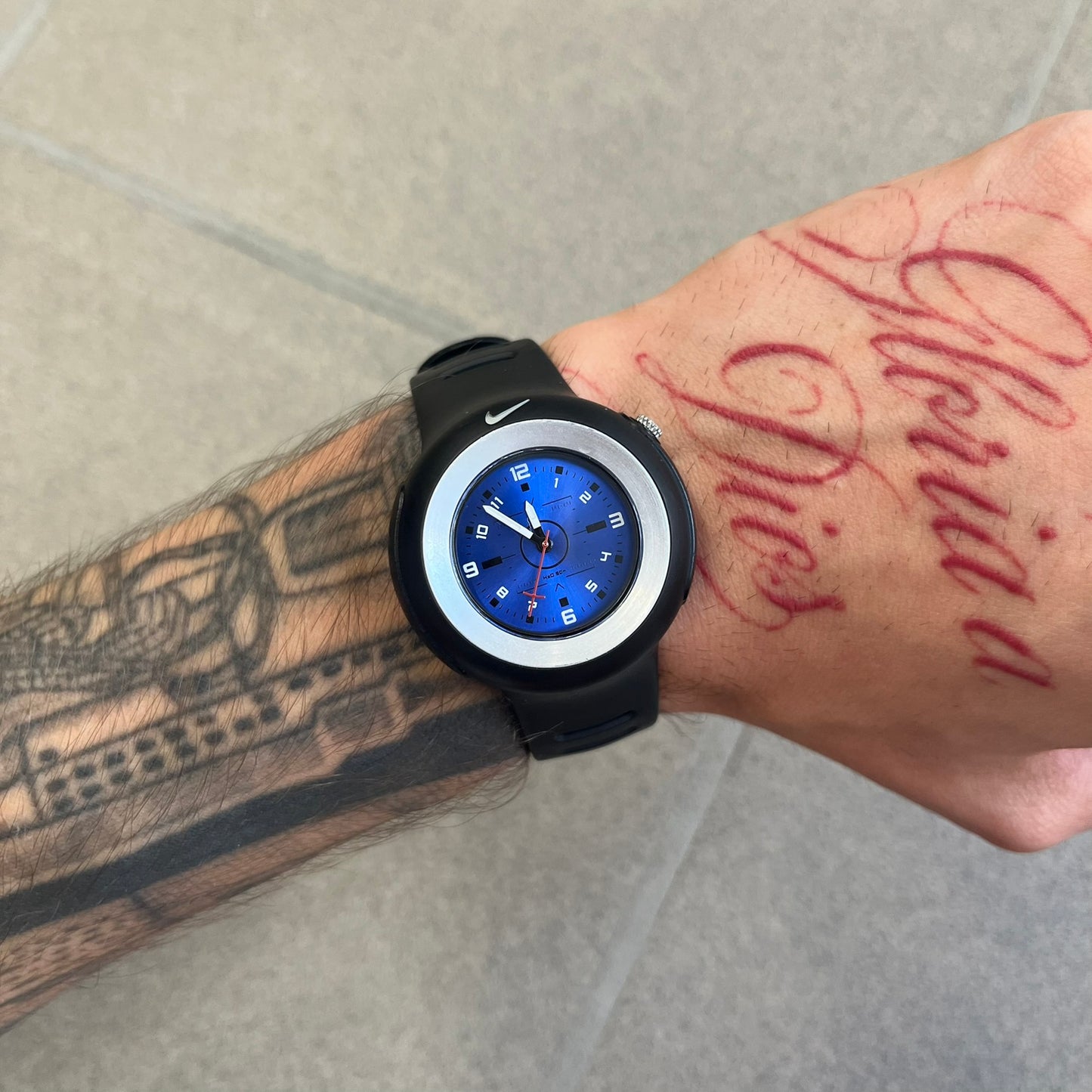 Nike Triax WK0009 Timing Range Blue Analog Watch features a black rubber strap and black rubber case with a silver-tone outline around the dial. The case has a grey Nike Swoosh at the top, while the metallic blue dial features white numerals and black hour markers.
