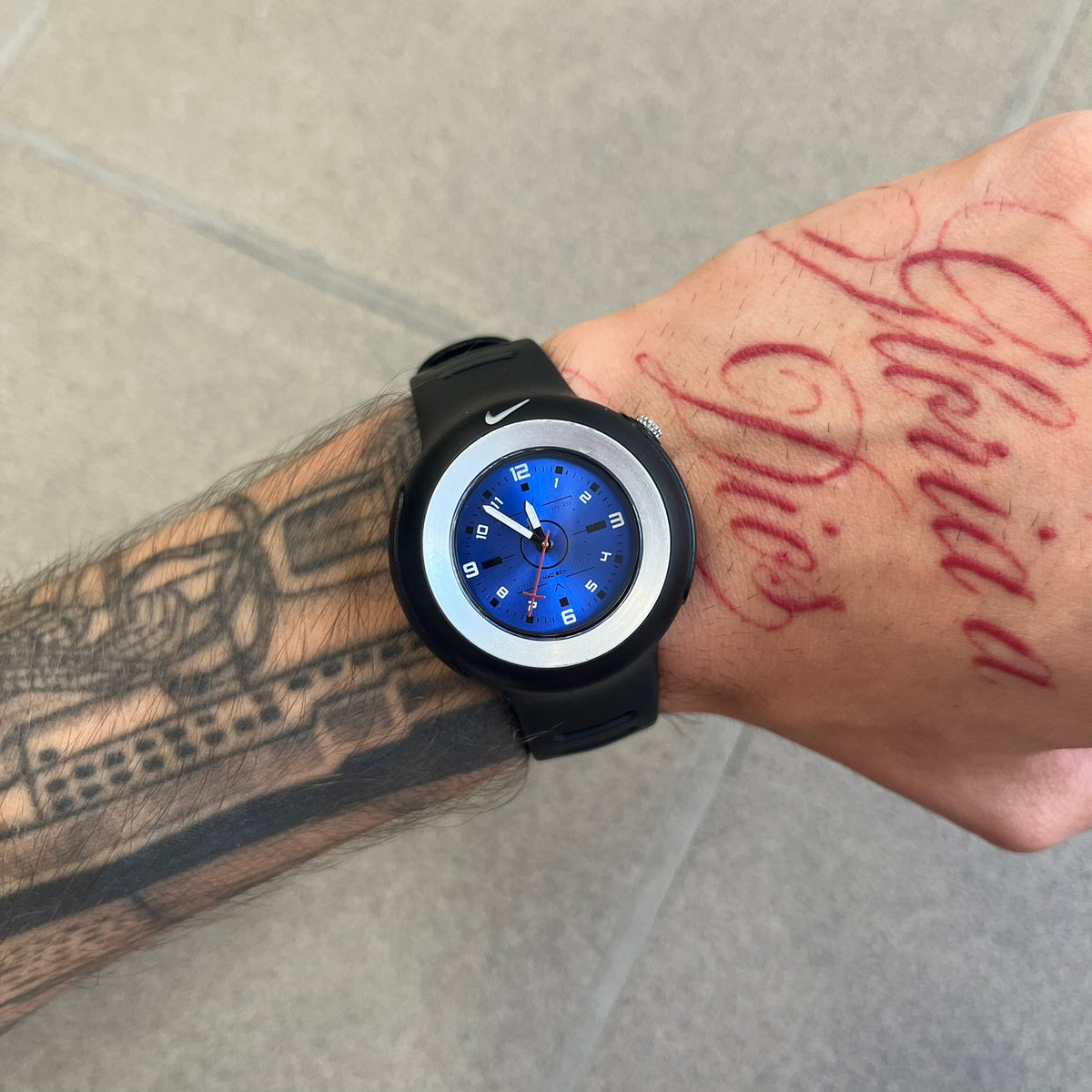 Nike Triax WK0009 Timing Range Blue Analog Watch features a black rubber strap and black rubber case with a silver-tone outline around the dial. The case has a grey Nike Swoosh at the top, while the metallic blue dial features white numerals and black hour markers.

