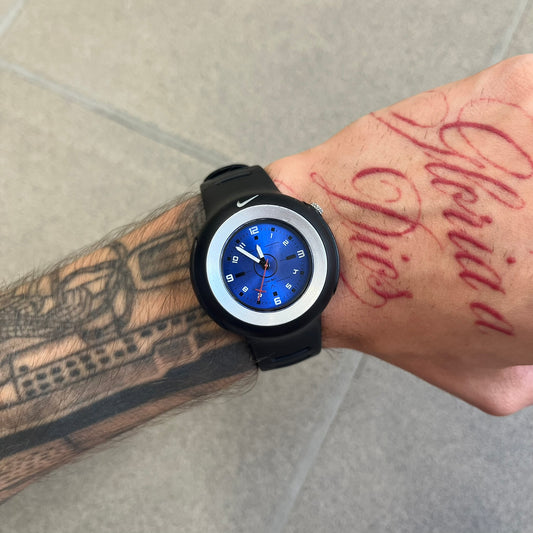 Nike Triax WK0009 Timing Range Blue Analog Watch features a black rubber strap and black rubber case with a silver-tone outline around the dial. The case has a grey Nike Swoosh at the top, while the metallic blue dial features white numerals and black hour markers.
