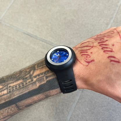 Nike Triax WK0009 Timing Range Blue Analog Watch features a black rubber strap and black rubber case with a silver-tone outline around the dial. The case has a grey Nike Swoosh at the top, while the metallic blue dial features white numerals and black hour markers.
