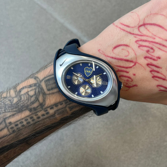 Nike Triax 'Boca Juniors' Swift 3i WR0091 Analog Watch with black rubber straps and a silver-tone case featuring a black Nike Swoosh at the top. The navy metallic dial has a recessed dotted texture, Boca Juniors crest at the top and three silver subdials for day, date, and hour. Seconds markings are printed on the inner edge of the case and are visible only at an angle.&nbsp;