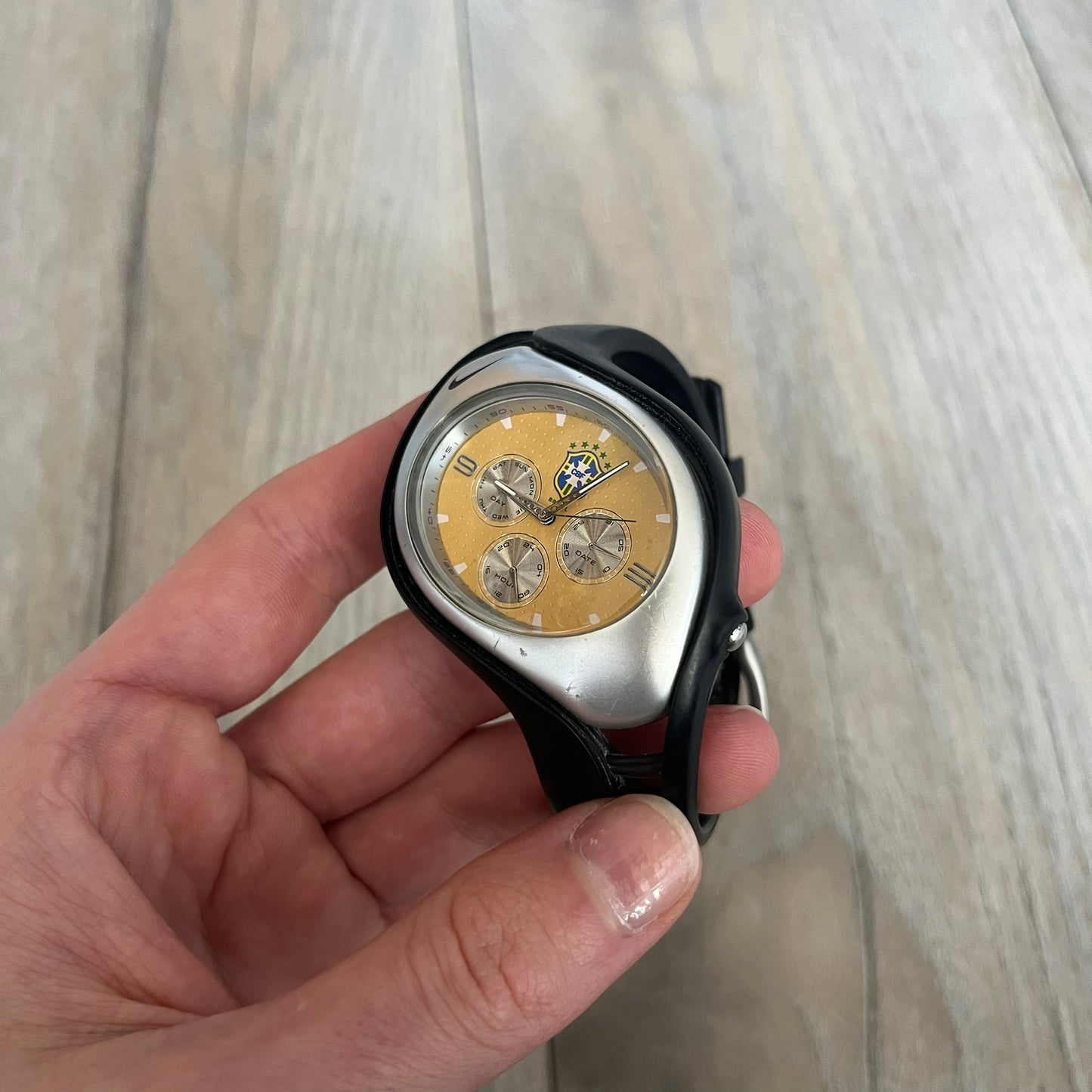 Nike Triax “Brasil” Swift 3i WR0091 analog watch with black rubber straps and a silver-tone case featuring a black Nike Swoosh. The yellow metallic dial has a recessed dotted texture, a Brazil crest at the top, three silver subdials for day, date, and hour, silver hands, and a green seconds hand. Seconds markings are printed on the inner edge of the case and are visible only at an angle. Sport-focused Triax case design with water-resistant construction.