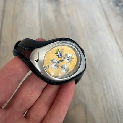 Nike Triax “Brasil” Swift 3i WR0091 analog watch with black rubber straps and a silver-tone case featuring a black Nike Swoosh. The yellow metallic dial has a recessed dotted texture, a Brazil crest at the top, three silver subdials for day, date, and hour, silver hands, and a green seconds hand. Seconds markings are printed on the inner edge of the case and are visible only at an angle. Sport-focused Triax case design with water-resistant construction.