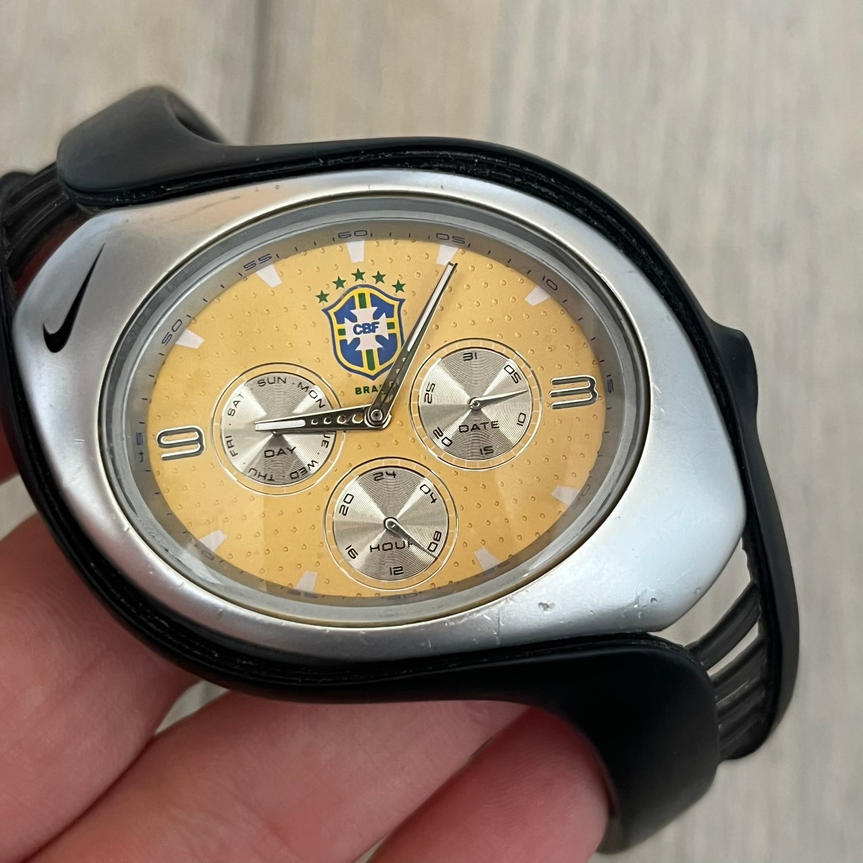 Nike Triax “Brasil” Swift 3i WR0091 analog watch with black rubber straps and a silver-tone case featuring a black Nike Swoosh. The yellow metallic dial has a recessed dotted texture, a Brazil crest at the top, three silver subdials for day, date, and hour, silver hands, and a green seconds hand. Seconds markings are printed on the inner edge of the case and are visible only at an angle. Sport-focused Triax case design with water-resistant construction.